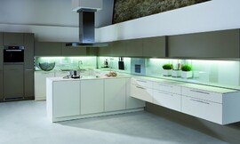 Deze keuken in F-vorm (!) valt op door het contrast tussen gebroken witte onderkasten, brons-bruine bovenkasten en een verlichte glazen keukenachterwand. Een gedeelte van de keukenwand zweeft boven de grond. Boven het centrale kookgedeelte hangt een opvallende afzuigkap. De keuken heeft een dun aanrechtblad dat optisch niet opvalt. Zuordnung: Stil Moderne keukens, Planungsart Detail keukenontwerp