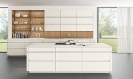 Een witte keuken met greeploze matte lakfronten die er vrijwel niet meer als keuken uitziet, doordat de apparatuur nauwelijks zichtbaar is. Dit maakt deze keuken met een hoog keukenblok aan de wand en een apart kookeiland uitermate geschikt om als open keuken naadloos aan te sluiten op de woonruimte. Ook de afwisseling tussen het tijdloze wit en het koperkleurige eiken uit het programma TIMBER zorgt voor een sfeervol en warm woonaccent. Zuordnung: Stil Design-keukens, Planungsart Open keuken (woonkeuken)