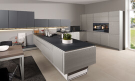 Natuurlijke houtstructuur in lichte en donkere grijstinten, spannend in scene gezet door metallic lak en de combinatie met matglas fronten. Ergonomisch is bovendien het in hoogte verstelbare kookeiland. Het extravagante oppervlakte van "Structura" ontstaat door diep te borstelen en aansluitend metallic lak aan te brengen.Tegen de muur staat het hoge kastenblok met in het midden op praktische hoogte de apparatuur. 

Haaks daarop, tegen de andere muur, staat het keukenblok met de spoelbak. Vanaf hier zijn er donkergrijze bovenkasten opgehangen, gaan over het kookeiland heen en lopen langs de muur verder waardoor de eetruimte of woonkamer  bij de keuken betrokken wordt. Zuordnung: Stil Moderne keukens, Planungsart Keuken met keukeneiland