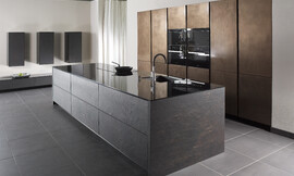 De open ontworpen, representatieve luxe keuken biedt maximale functionaliteit met veel bergruimte. Voor het kookeiliand is gebruik gemaakt van echte grijze leisteen. Het dunne, hoogglanzende aanrechtblad zorgt voor een interessant contrast. Op het kookeiland zijn de spoelbak en het fornuis opgenomen. In de wand zijn in hoge kasten de apparaten opgenomen, twee daarvan op een praktische werkhoogte. De luxe en exclusieve uitstraling van deze greeploze keuken wordt door niets verstoord. Zuordnung: Stil Luxe keukens, Planungsart Keuken met keukeneiland