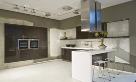 Deze moderne keuken leunt optisch op het contrast tussen sepia bruine kasten en een wit aanrechtblad, bar en achterwand. De apparatuurkast links is zwevend gemonteerd tegen een grijze strook. Zuordnung: Stil Moderne keukens, Planungsart Open keuken (woonkeuken)