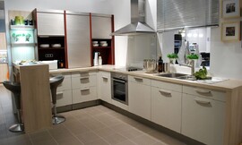 Deze gezellige L-vormige keuken biedt zeven onderkasten met beige structuurlak. Op het stompe gedeelte van de L-vorm zijn drie opzetkasten geplaatst die voorzien zijn van praktische rolluiken. Aan dit gedeelte sluit een bar in houtoptiek aan, zodat hier een zitgelegenheid voor twee personen wordt geboden. Het fornuis, de oven en de spoelbak zijn opgnomen in het langere gedeelte van de L-vorm. Zuordnung: Stil Design-keukens, Planungsart Keuken met keukeneiland