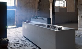 Kubistisch gevormd keukenblok. Platte, greeploze fronten met subtiel aanrechtblad in dezelfde grijstint. Keukenkraan komt uit een achterwand. Gootsteen afdekbaar. Keuken op sokkel. Zuordnung: Stil Design-keukens, Planungsart Open keuken (woonkeuken)