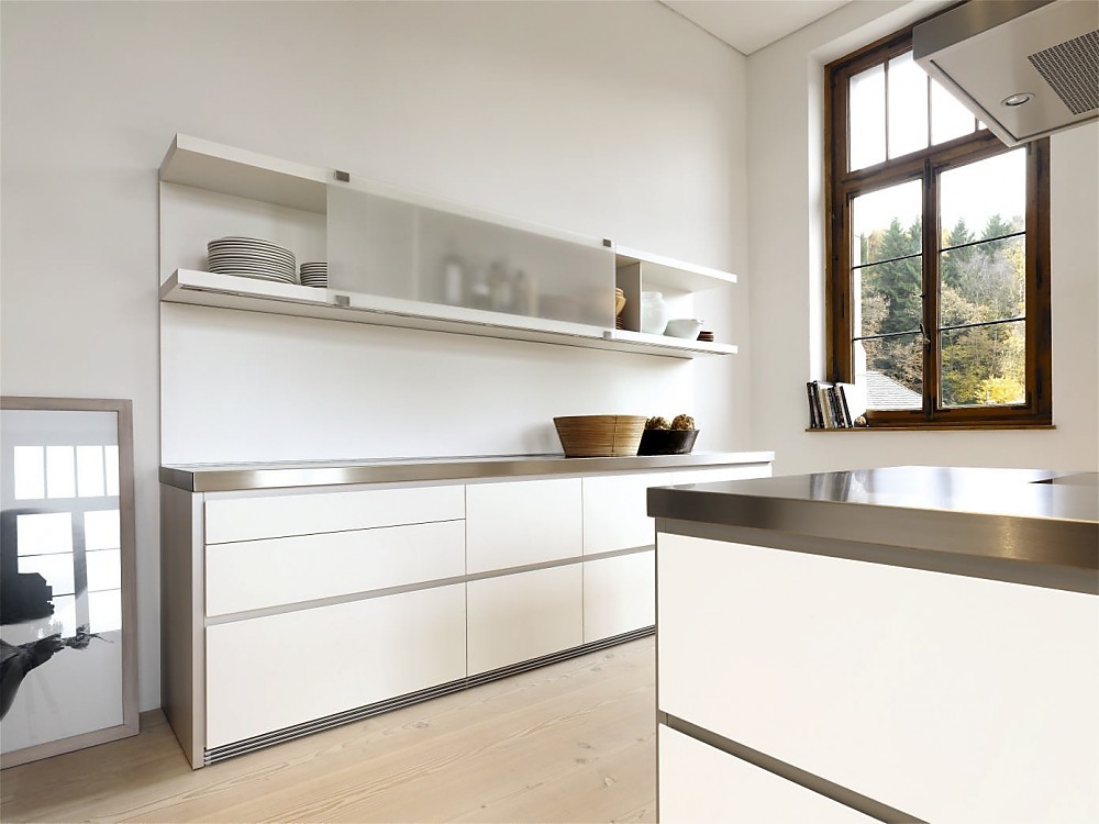 Deze moderne witte designkeuken maakt met zijn roestvrijstalen aanrechtbladen en greeploze witte fronten een strakke indruk. Aan de wand is een keukenblok geplaatst met grote, praktische lades. De lage kast daarboven heeft verschuifbare matglazen fronten en is voor een deel open. Het kookeiland heeft hetzelfde design: roestvrijstalen werkblad, witte greeploze fronten. Niets verstoort hier de kalmte die deze keuken uitstraalt. Zuordnung: Stil Design-keukens, Planungsart Keuken met keukeneiland