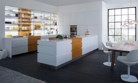 In deze moderne U-vormige keuken ziet u wat voor een effect bereikt kan worden door kleurcontrasten te introduceren in verder identieke keukenmeubels. Het stralende wit in de twee tegenover elkaar staande keukenblokken wordt afgewisseld met de trendy kleur "curcuma, maar ook de kleuren "saffraan" en "petrol" zijn momenteel verkrijgbaar. Het tegen de muur staande keukenblok heeft over de hele lengte open kasten vanaf het aanrechtblad naar boven. Perfect om mooie decoratieve accenten te zetten en praktisch omdat u alles meteen bij de hand heeft. In het andere keukenblok dat daar tegenover ligt bevindt zich het fornuis. Dit keukenblok accentueert het open karakter van de keuken, doordat er geen kasten boven zijn aangebracht. Zo kunt u tijdens het koken contact houden met de anderen die aan de keukentafel zitten. Het "kookschiereiland" en het keukenblok met de spoelbak zijn met elkaar verbonden door een wand met greeploze witte kasten onderbroken in het midden door apparatuur op praktische ooghoogte. Zuordnung: Stil Design-keukens, Planungsart Open keuken (woonkeuken)