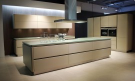 Ruime eilandkeuken met kookveld op het kookeiland. Beige fronten met greeplijsten, afgewerkt met rondingen. Dun glazen aanrechtblad, kleurtint richting groen. Zuordnung: Stil Design-keukens, Planungsart Keuken met zitgelegenheid