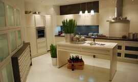 Deze keuken laat u zien wat u met een relatief grote ruimte kunt doen. Verschillende keukenmeubels tegen de wanden en een keukeneiland in het midden. Crèmetinten domineren deze keuken. Het aanrechtblad heeft een donkerdere tint. De fronten hebben brede grepen voor een iets traditionelere look. De kast links heeft glazen deurtjes. Zuordnung: Stil Moderne keukens, Planungsart Keuken met keukeneiland