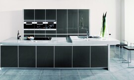 Elegantie en rechtlijnigheid in een donkere metallic tint achter glas met net aluminium kader. Hier ziet u ook het bijzondere effect van een aanrechtblad met verschillende hoogten. Rechts ziet u het verhoogde deel met de spoelbak en de eetbar. Zuordnung: Stil Moderne keukens, Planungsart Open keuken (woonkeuken)