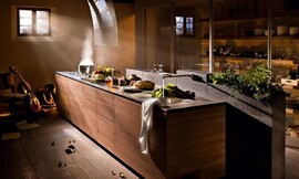 Deze greeploze keuken buit de esthetische kracht van hout volledig uit voor een symbiose tussen design en klassieke accenten. Door het greeploze ontwerp komt het hout maximaal tot zijn recht. De keukenkraan komt uit een achterwand van donkere steenblokken, die net iets hoger dan de keukenkastjes is en nog plaats biedt aan diverse kruidenplantjes. De design-afzuigkap met holle glasplaat maakt de keuken af. Zuordnung: Stil Design-keukens, Planungsart Keuken met keukeneiland