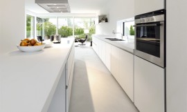 Witte design keuken, hier gefotografeerd vanaf de zithoek aan het kookeiland. De fronten zijn greeploos; hier ziet u ook goed hoe de fronten schuin afgerond zijn zodat deze makkelijk te openen zijn. Een kookeiland kan een relatief smalle ruimte goed in verschillende werkbereiken indelen. Zuordnung: Stil Design-keukens, Planungsart Keuken met keukeneiland