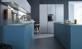Deze uit drie blokken bestaande keuken met kookeiland bestaat uit mat glas en greeploze fronten met hoogglans lak. De kasten van het kookeiland, vijf stuk in totaal, werken kwadratisch in fris gedempt blauw, de kleur die ook doorloopt in het werkblad. Ook de kasten die daar tegenover staan zetten de frisse blauwe tint voort. Daarboven hangen langwerpige, maar lage kasten met een lichtgrijze hoogglans lak. Haaks op de beide keukenblokken staat het lichtgrijze keukenelement met twee hoge kasten met daartussen een onderkast, de roestvrijstalen oven en een bovenkast. Mat glas (gedempt blauw) en hoogglans lak (lichtgrijs) zijn de homogene tinten van deze designkeuken. Zuordnung: Stil Moderne keukens, Planungsart U-vormige keuken