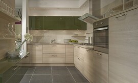 Bovenkasten met olijfgroene hoogglans gelakte fronten. Daaronder licht houten keukenmeubelen met een even licht zandkleurig aanrechtblad. Voor de keukenachterwand tussen keukenblok en bovenkast is geribbeld glas gebruikt. Zuordnung: Stil Moderne keukens, Planungsart U-vormige keuken