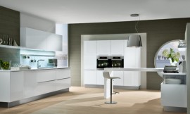 Een symmetrische apparaathoogkast met twee apparaten, een keukenblok met grote lades/fronten en een bartafel die ver de kamer in reikt. Het kookveld is op de bartafel aangebracht met daarboven een afzuigkap met lampenkap. Dit alles in stralend wit. Zuordnung: Stil Moderne keukens, Planungsart Keuken met zitgelegenheid