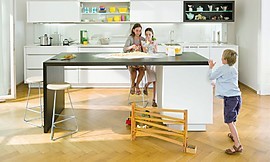 Deze familiekeuken bestaat uit een breed keukenblok met hangkasten en een simpel kookeiland met zitgelegenheid. Witte fronten met brede greeplijst. Bovenkast met zich naar boven openende kleppen. Zuordnung: Stil Luxe keukens, Planungsart keukenblok