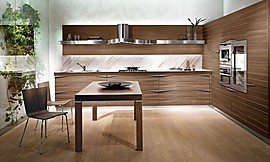 L-keuken met walnoot hout. Deze kleurtint vindt u in de keukenmeubels, tafel en stoelen. Het belangrijkste contrast is met het metaal van de afzuiging en de andere keukenapparatuur. Horizontale patronen in het hout en stijlvolle grepen maken het ontwerp af. Zuordnung: Stil Moderne keukens, Planungsart L-vormige keuken