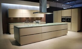 Ruime eilandkeuken met kookveld op het kookeiland. Beige fronten met greeplijsten, afgewerkt met rondingen. Dun glazen aanrechtblad, kleurtint richting groen. Zuordnung: Stil Design-keukens, Planungsart Open keuken (woonkeuken)