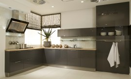 Deze moderne L-vormige keuken heeft donkere antraciet fronten (hoogglans lak) met brede grepen. Deze contrasteren met een zandkleurig aanrechtblad. Het fornuis bevindt zich aan de ene kant van de keuken, met daaronder handige grote lades en de spoelbak bevindt zich aan de andere kant. Boven de spoelbak zijn drie hangkastjes bevestigd. Daaronder is een glazen legplank aan de muur geplaatst. Ideaal om decoratief te gebruiken, of tegelijkertijd handig met mooie glazen. De keuken wordt afgesloten door hoge kasten met op ooghoogte een open gedeelte. De afzuigkap valt door zijn bijzondere design zeker op. Zuordnung: Stil Moderne keukens, Planungsart Keuken met keukeneiland