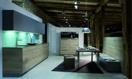 Deze design keuken in L-vorm combineert licht hout (zand) voor de greeploze fronten van de onderkast met donker/paars-blauwe fronten van de bovenkasten. Het aanrechtblad heeft een donkerdere bruintint en loopt als sideboard om de kast heen. Een verlichte glazen plaat geeft de keuken een fonkelend randje. Zuordnung: Stil Design-keukens, Planungsart keukenblok
