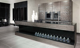 Moderne eilandkeuken met een werkblad van roestvrij staal op sterk geborsteld fineer. In combinatie met glanzend zwart achter glas een echte eye catcher. Gekookt wordt er op het kookeiland, ook de spoelbak bevindt zich op het eiland. Het kookeiland heeft aan de voorkant boven en onder een strook met het geborstelde fineer. Daartussen is een inkeping van het glanzend zwarte materiaal. Geschikt voor o.a. decoartieve doeleinden. Tegen de muur staan de kasten die veel bergruimte opleveren en in het midden van die kasten is de apparatuur op makkelijke bedienhoogte aangebracht. Zuordnung: Stil Moderne keukens, Planungsart Keuken met keukeneiland