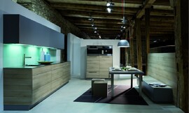 Deze design keuken in L-vorm combineert licht hout (zand) voor de greeploze fronten van de onderkast met donker/paars-blauwe fronten van de bovenkasten. Het aanrechtblad heeft een donkerdere bruintint en loopt als sideboard om de kast heen. Een verlichte glazen plaat geeft de keuken een fonkelend randje. Zuordnung: Stil Moderne keukens, Planungsart Keuken met keukeneiland