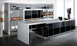 Moderne designkeuken met licht grijs betegeld aanrechtblad en wangen. De kasten zijn voorzien van hoogglans zwarte fronten. Het aanrechtblad is als het ware op de kasten gelegd en is daardoor iets hoger. Bovendien is er afstand bewaard ten opzichte van de kasten, zodat er een fraaie zitgelegenheid is ontstaan.

Het wandelement is betegeld met dezelfde tegels als die op het kookeiland gebruikt zijn. De kasten die in het wandelement opgenomen zijn hebben ook hier dezelfde zwarte hoogglans fronten. De apparatuur is op praktische werkhoogte naast elkaar aangebracht. Als extra luxe-element dient het butchter-block te worden genoemd, aangebracht naast de kasten in het wandelement. Daarboven bevindt zich de mogelijkheid de wijnverzameling op rekken te bewaren. Zuordnung: Stil Design-keukens, Planungsart Keuken met zitgelegenheid