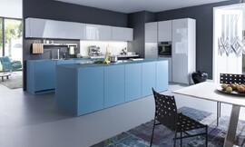 Deze uit drie blokken bestaande keuken met kookeiland bestaat uit mat glas en greeploze fronten met hoogglans lak. De kasten van het kookeiland, vijf stuk in totaal, werken kwadratisch in fris gedempt blauw, de kleur die ook doorloopt in het werkblad. Ook de kasten die daar tegenover staan zetten de frisse blauwe tint voort. Daarboven hangen langwerpige, maar lage kasten met een lichtgrijze hoogglans lak. Haaks op de beide keukenblokken staat het lichtgrijze keukenelement met twee hoge kasten met daartussen een onderkast, de roestvrijstalen oven en een bovenkast.  
Mat glas (gedempt blauw) en hoogglans lak (lichtgrijs) zijn de homogene tinten van deze designkeuken. De meubels werken kwadratisch en fris door greeploze fronten en een aanrechtblad in dezelfde tint. Zie ook het bijzondere effect van de combinatie van een design keuken met een vloerkleed. Zuordnung: Stil Design-keukens, Planungsart L-vormige keuken