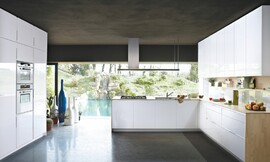 Witte keuken in U-vorm met greeplijsten en licht-houten sideboards en aanrechtbladen. Kookbereik en gootsteen midden in de keuken. Met een stijlvolle afzuigkap. Tussen de onderkasten en hoogkasten (rechts) ontstaat een nis die extra opbergruimte biedt. Zuordnung: Stil Moderne keukens, Planungsart Open keuken (woonkeuken)