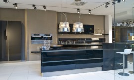 Deze moderne zwarte eilandkeuken in hoogglans uitvoering biedt behalve het kookeiland een keukenblok aan de wand met de spoelbak, onder- en bovenkasten. De bar aan het kookeiland biedt zitgelegenheid voor twee personen. De apparatuur is op praktische werkhoogte in de wand opgenomen. De koel- vriescombinatie is in separaat geplaatste hoge kasten geplaatst. De greeploze zwarte hoogglans fronten reflecteren licht en het patroon van de vloer. Hier ziet u ook een dubbele afzuigkap in een lampenkap. Zuordnung: Stil Moderne keukens, Planungsart Keuken met keukeneiland