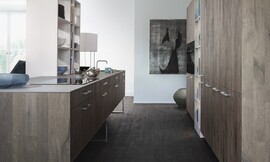 Knoesten, scheuren en het geborstelde oppervlak geven de eikenfronten "Xylo" een knoestig karakter. Het keukeneiland met gootsteen zweeft grotendeels boven de vloer. De keuken gaat harmonisch over in de woonkamer. Zuordnung: Stil Design-keukens, Planungsart Keuken met keukeneiland