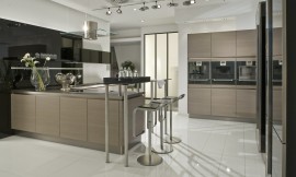 De apparatuur is in deze moderne keuken met greeploze eiken houten fronten op praktische werkhoogte in hoge kasten aan de wand opgenomen. Levendig en modern wordt de keuken door het wandpaneel dat achter het L-vormige keukenblok is bevestigd: dit is namelijk hoogglanzend zwart en biedt de mogelijkheid legplanken op te nemen. Het L-vormige gedeelte van de keuken bestaat allereerst uit een blok dat aan de wand is geplaatst, met spoelbak en daarboven lage hoogglanzende zwarte bovenkasten. Om de hoek is dan het kookeiland waarop het fornuis is bevestigd, daarboven hangt de moderne afzuigkap van roestvrij staal en glas. Aan het eind van het kookeiland is een bar opgenomen die aan de ene kant op twee korte, ronde metalen poten op het aanrechtblad rust en aan de andere kant op twee hoge poten staat. De zitgelegenheid die hierdoor is ontstaan biedt plaats aan twee personen. Zuordnung: Stil Moderne keukens, Planungsart U-vormige keuken