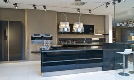 Deze moderne zwarte eilandkeuken in hoogglans uitvoering biedt behalve het kookeiland een keukenblok aan de wand met de spoelbak, onder- en bovenkasten. De bar aan het kookeiland biedt zitgelegenheid voor twee personen. De apparatuur is op praktische werkhoogte in de wand opgenomen. De koel- vriescombinatie is in separaat geplaatste hoge kasten geplaatst. De greeploze zwarte hoogglans fronten reflecteren licht en het patroon van de vloer. Hier ziet u ook een dubbele afzuigkap in een lampenkap. Zuordnung: Stil Moderne keukens, Planungsart Keuken met keukeneiland