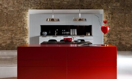 Een simpel, symmetrisch maar elegant keukenblok tegen de wand wordt afgemaakt met een afgestemde tafel en rood kastmeubel. De buitenste rij kasten is greeploos en de lades in het midden hebben horizontale grepen. Simpele witte fronten contrasteren met de felrode kast in de voorgrond. Zuordnung: Stil Luxe keukens, Planungsart Keuken met keukeneiland