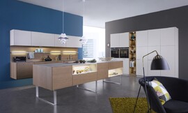 De combinatie van bijzonder natuurlijk werkende kopereiken fronten met matte sahara-lak zorgt voor een interessante spanning. In de hangkast is een efficiënte afzuigkap geïntegreerd die geen luchtafvoer behoeft. De keuken bestaat uit drie delen: het opgehangen keukenblok met fornuis en spoelbak, de eikenhouten fronten en daarboven de witte hangkasten; het keukeneiland in het midden, staande op metalen open poten die het geheel daardoor een luchtig karakter geven en de hoge witte kasten aan de andere wand waarin op praktische werkhoogte de roestvrijstalen apparatuur is aangebracht. Zuordnung: Stil Moderne keukens, Planungsart U-vormige keuken