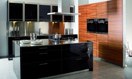 Deze keuken bestaat uit twee keukenblokken die tegen de wand geplaatst zijn en een apart kookeiland. In een van de twee keukenblokken met het natuurlijke tineo fineer, met duidelijke houtstructuur, is de apparatuur op praktische werkhoogte opgenomen. Het kookeiland en het andere keukenblok zijn voorzien van een in 13 stappen vervaardigde zwarte hoogglans lak, waardoor een spannend contrast met de houtoptiek van de hoge apparatuurkasten is ontstaan. Het roestvrij staal van de sokkels komt weer terug in de vijf hangkasten boven de spoelbak, die samen met de onderkasten en de grote lades voor voldoende bergruimte zorgen. Zuordnung: Stil Design-keukens, Planungsart Keuken met keukeneiland