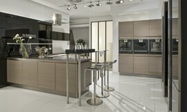 De apparatuur is in deze moderne keuken met greeploze eiken houten fronten op praktische werkhoogte in hoge kasten aan de wand opgenomen. Levendig en modern wordt de keuken door het wandpaneel dat achter het L-vormige keukenblok is bevestigd: dit is namelijk hoogglanzend zwart en biedt de mogelijkheid legplanken op te nemen. Het L-vormige gedeelte van de keuken bestaat allereerst uit een blok dat aan de wand is geplaatst, met spoelbak en daarboven lage hoogglanzende zwarte bovenkasten. Om de hoek is dan het kookeiland waarop het fornuis is bevestigd, daarboven hangt de moderne afzuigkap van roestvrij staal en glas. Aan het eind van het kookeiland is een bar opgenomen die aan de ene kant op twee korte, ronde metalen poten op het aanrechtblad rust en aan de andere kant op twee hoge poten staat. De zitgelegenheid die hierdoor is ontstaan biedt plaats aan twee personen. Zuordnung: Stil Moderne keukens, Planungsart Detail keukenontwerp