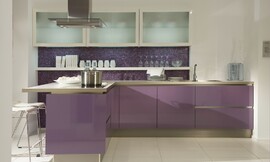 Deze L-vormige keuken leunt op het subtiele contrast tussen het ongebroken paars van de greeploze fronten en het iets donkerdere paars van de mozaïek achterwand. Daarnaast heeft deze glazen bovenkasten, hoge sokkels en wit aanrechtblad waar aan gezeten kan worden. Zuordnung: Stil Moderne keukens, Planungsart keukenblok