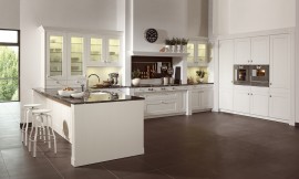 Deze witte keuken laat de moderne interpretatie van een landelijke keuken zien. Heldere lijnvoering, lichte tinten en verfijnde houtoppervlakten. De keuken bestaat uit drie delen: Tegen de wand zijn hoge kasten geplaatst waarin op praktische werkhoogte de roestvrijstalen apparatuur is opgenomen. Tegen de andere wand is een luxe fornuis in een schouw geplaatst, wat bijdraagt aan de landelijke en klassieke uitstraling van de keuken. Naast de schouw zijn aan allebei de kanten twee onderkasten met daarboven twee hangkasten met glazen deurtjes opgenomen. Links sluit een hoge opzetkast aan met dezelfde glazen deuren. Deze opzetkast is geplaatst op het keukeneiland. De spoelbak bevindt zich op het schiereiland. Aan de andere kant van het eiland is door het fraaie zwarte werkblad dat iets uitsteekt een ruime zitgelegenheid ontstaan. Al met al een keuken die aan alle moderne eisen voldoet en desondanks door de schouw en de cassettenfronten een klassieke en landelijke charme biedt. Zuordnung: Stil Landelijke keukens, Planungsart Keuken met keukeneiland