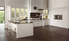 Deze witte keuken laat de moderne interpretatie van een landelijke keuken zien. Heldere lijnvoering, lichte tinten en verfijnde houtoppervlakten. De keuken bestaat uit drie delen: Tegen de wand zijn hoge kasten geplaatst waarin op praktische werkhoogte de roestvrijstalen apparatuur is opgenomen. Tegen de andere wand is een luxe fornuis in een schouw geplaatst, wat bijdraagt aan de landelijke en klassieke uitstraling van de keuken. Naast de schouw zijn aan allebei de kanten twee onderkasten met daarboven twee hangkasten met glazen deurtjes opgenomen. Links sluit een hoge opzetkast aan met dezelfde glazen deuren. Deze opzetkast is geplaatst op het keukeneiland. De spoelbak bevindt zich op het schiereiland. Aan de andere kant van het eiland is door het fraaie zwarte werkblad dat iets uitsteekt een ruime zitgelegenheid ontstaan. Al met al een keuken die aan alle moderne eisen voldoet en desondanks door de schouw en de cassettenfronten een klassieke en landelijke charme biedt. Zuordnung: Stil Landelijke keukens, Planungsart Open keuken (woonkeuken)