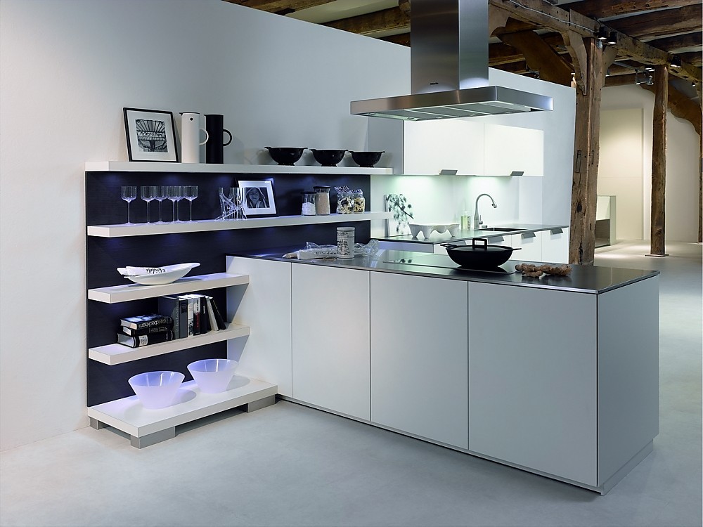 Deze open keuken heeft een gereduceerde look met witte fronten en een dun, metallic aanrechtblad. Het kookeiland staat haaks op een open wandgedeelte. Deze open wand met legplanken is ideaal om alles direct bij de hand te hebben, maar kan tegelijkertijd ook decoratief worden gebruikt. De blauwe achtergrond van de open kast zorgt voor een speels accent. Op deze open wandkast sluit het keukenblok met de spoelbak aan. De onder- en bovenkasten bieden voldoende bergruimte. Een keukenplanning zoals deze is ook erg geschikt voor grote ruimtes die deels als woonkamer functioneren. Zuordnung: Stil Design-keukens, Planungsart Open keuken (woonkeuken)