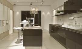 Keuken met indrukwekkende hoogglans fronten en keukeneiland met wit aanrechtblad en hoge bar met zitgelegenheid op barkrukken. De grote, ongebroken sepia bruine vlakken van de greeploze fronten geven deze keuken een krachtig timbre. Zuordnung: Stil Moderne keukens, Planungsart keukenblok