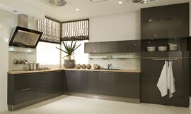 Deze moderne L-vormige keuken heeft donkere antraciet fronten (hoogglans lak) met brede grepen. Deze contrasteren met een zandkleurig aanrechtblad. Het fornuis bevindt zich aan de ene kant van de keuken, met daaronder handige grote lades en de spoelbak bevindt zich aan de andere kant. Boven de spoelbak zijn drie hangkastjes bevestigd. Daaronder is een glazen legplank aan de muur geplaatst. Ideaal om decoratief te gebruiken, of tegelijkertijd handig met mooie glazen. De keuken wordt afgesloten door hoge kasten met op ooghoogte een open gedeelte. De afzuigkap valt door zijn bijzondere design zeker op. Zuordnung: Stil Moderne keukens, Planungsart Detail keukenontwerp