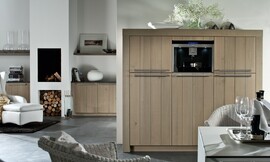 Hier ziet u een detailfoto van de hoogkast/apparatuurkast met brede wangen, stanggrepen en verticale strepen in het hout. Ook ziet u hoe hetzelfde concept is doorgevoerd in de woonkamer. Aparte eettafel met rieten stoelen. Zuordnung: Stil Landelijke keukens, Planungsart Keuken met keukeneiland
