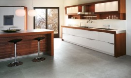 Deze designkeuken valt op door de combinatie van brede, donkere houten segmenten met heldere witte vlakken (fronten). Het donkere wandelement bevat zowel spoelbak als fornuis en handige witte lades. Daarboven een bijzonder hanggedeelte met twee gesloten kasten waar mat glazen deurtjes voor zitten, daaronder een legplank. Tussen de twee bovenkasten is de zeer bijzondere afzuigkap met een golvende vorm opgehangen. Het extra keukeneiland biedt een bar van hetzelfde hout als de wangen van het wandelement, waaraan gezellig gegeten of geborreld kan worden. Zuordnung: Stil Moderne keukens, Planungsart Keuken met keukeneiland