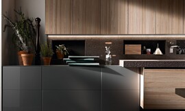 Hier ziet u hoe de optiek van het gebeitste aanrechtblad doorgezet wordt in de keukenachterwand. Een horizontale verlichtingsstrook benadrukt het reliëf van dit materiaal. Zuordnung: Stil Design-keukens, Planungsart Open keuken (woonkeuken)