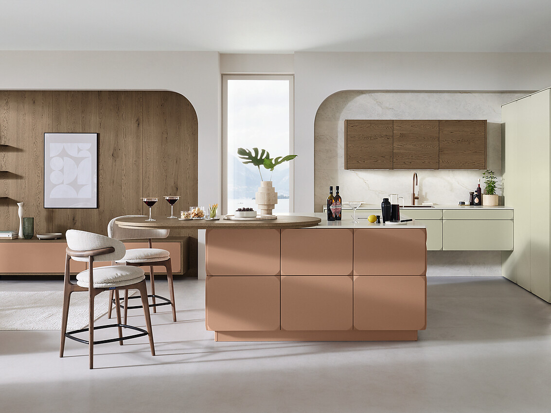 Mat gelakte fronten in warm terracotta met afgeschuinde hoeken in retrodesign ontmoeten saliegroene bovenkasten en een donkere eikenlook. Het ovale houten tafelblad sluit naadloos aan op het eiland en verbindt koken, eten en wonen tot een harmonieus geheel. Zuordnung: Stil Moderne keukens, Planungsart Keuken met keukeneiland