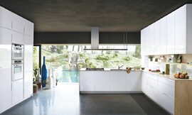 Witte keuken in U-vorm met greeplijsten en licht-houten sideboards en aanrechtbladen. Kookbereik en gootsteen midden in de keuken. Met een stijlvolle afzuigkap. Tussen de onderkasten en hoogkasten (rechts) ontstaat een nis die extra opbergruimte biedt. Zuordnung: Stil Moderne keukens, Planungsart Open keuken (woonkeuken)
