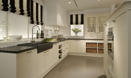 Landelijke L-vormige keuken met moderne touch. Hier zijn de witte cassettefronten gecombineerd met een contrasterend zwart aanrechtblad. De spoelbak heeft dezelfde kleur, maar is iets hoger dan het aanrechtblad geplaatst. In hetzelfde keukenblok als de spoelbak is ook het fornuis opgenomen. In dit deel van de keuken zijn er alleen onderkasten, voor een deel met praktische grote lades, een ander deel met deurtjes en er is ook een praktisch wijnrek beschikbaar voor de wijnliefhebber. Om de hoek is er eerst een onderkast met twee deurtjes, daarna twee open onderkasten met manden, die het geheel een landelijke sfeer geven. Op het aanrechtblad daar boven zijn twee hoge kasten met fraaie sierlijsten gezet. De glazen deurtjes van de twee kasten maken het mogelijk een blik te werpen op het mooie servies, de bijzondere wijnglazen of andere interessante keukenaccessoires. Onder de kast met glazen deuren zijn er nog vier handige lades aangebracht. Als los element is er een apparatuurkast geplaatst. Ook deze kast wordt aan de bovenkant gesierd door een mooie afsluitende lijst. In het midden van dit keukenelement bevindt zich op handige werkhoogte de oven. Direct daaronder is er een open kast met legplank. Zuordnung: Stil Landelijke keukens, Planungsart U-vormige keuken