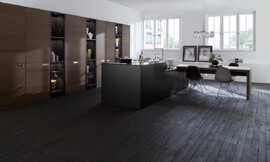 Vrijwel niet meer te herkennen als keuken, doordat o.a. de apparatuur achter hoge houten schuifdeuren in de wand is aangebracht. Het kookeiland is strak vormgegeven, zwart van kleur en zonder enige tierelantijnen. Het fornuis en de spoelbak met mengkraan vallen nauwelijks op. Aan één kant is het kookeiland iets lager gemaakt. Daarop ligt het hout voor een lange eettafel. Al met al een terughoudende keuken die prima in een woonruimte past. Zuordnung: Stil Design-keukens, Planungsart Keuken met zitgelegenheid