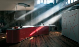 Dit vrijstaande in een lichte boog vormgegeven keukenblok is het hart van de moderne greeploze keuken. Op dit rode keukeneiland bevinden zich zowel de spoelbak als het fornuis. Daarboven is de glazen design-afzuigkap aangebracht. De bartafel die aan het eiland is bevestigd zorgt voor een hoge zitgelegenheid. Tegen de wand zijn hoge kasten o.a. voor de koelapparatuur geplaatst. De deuren van deze kasten zijn een modern kunstwerk op zich. Het heldere rood van het keukenblok komt hierin weer terug. Zuordnung: Stil Luxe keukens, Planungsart Keuken met keukeneiland