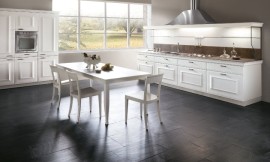 L-keuken in landelijke stijl met cassettefronten. Witte meubels met grijs werkblad. Zitgelegenheid aan vrijstaande tafel. Boven het werkblad is een schouw met ingebouwde afzuigkap aangebracht, die bijdraagt aan het symmetrische karakter van het meubel. Zuordnung: Stil Landelijke keukens, Planungsart Keuken met keukeneiland